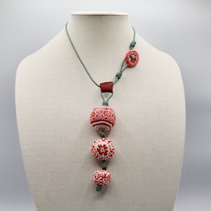 May include: A green cord necklace with a red and white patterned ceramic pendant. The pendant is made up of three round beads with intricate floral designs.