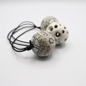 May include: A black leather cord necklace with three white and gray patterned ceramic beads. The beads have intricate designs and are connected with a knot.