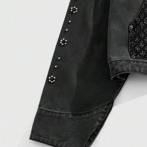 May include: Close-up of a black denim jacket sleeve. The sleeve is adorned with small, silver-colored floral embellishments and silver studs. The jacket has a classic design with a buttoned cuff and a split hem.