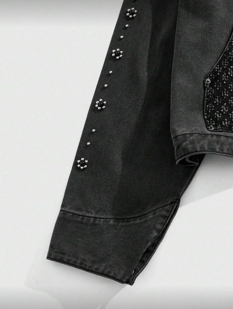 May include: Close-up of a black denim jacket sleeve. The sleeve is adorned with small, silver-colored floral embellishments and silver studs. The jacket has a classic design with a buttoned cuff and a split hem.