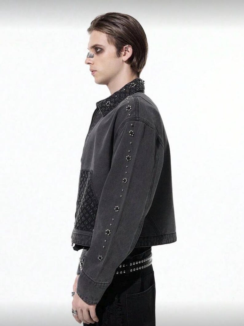 May include: A dark gray denim jacket with decorative studs along the sleeves and collar. The jacket features a textured panel on the front and a studded belt. The jacket is a stylish piece of outerwear.