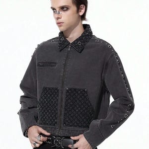 May include: A dark gray denim jacket with a zip-up front, embellished with studs on the collar, sleeves, and pockets. The jacket features a textured pattern on the pockets and collar. The model is wearing a black belt with a decorative buckle.