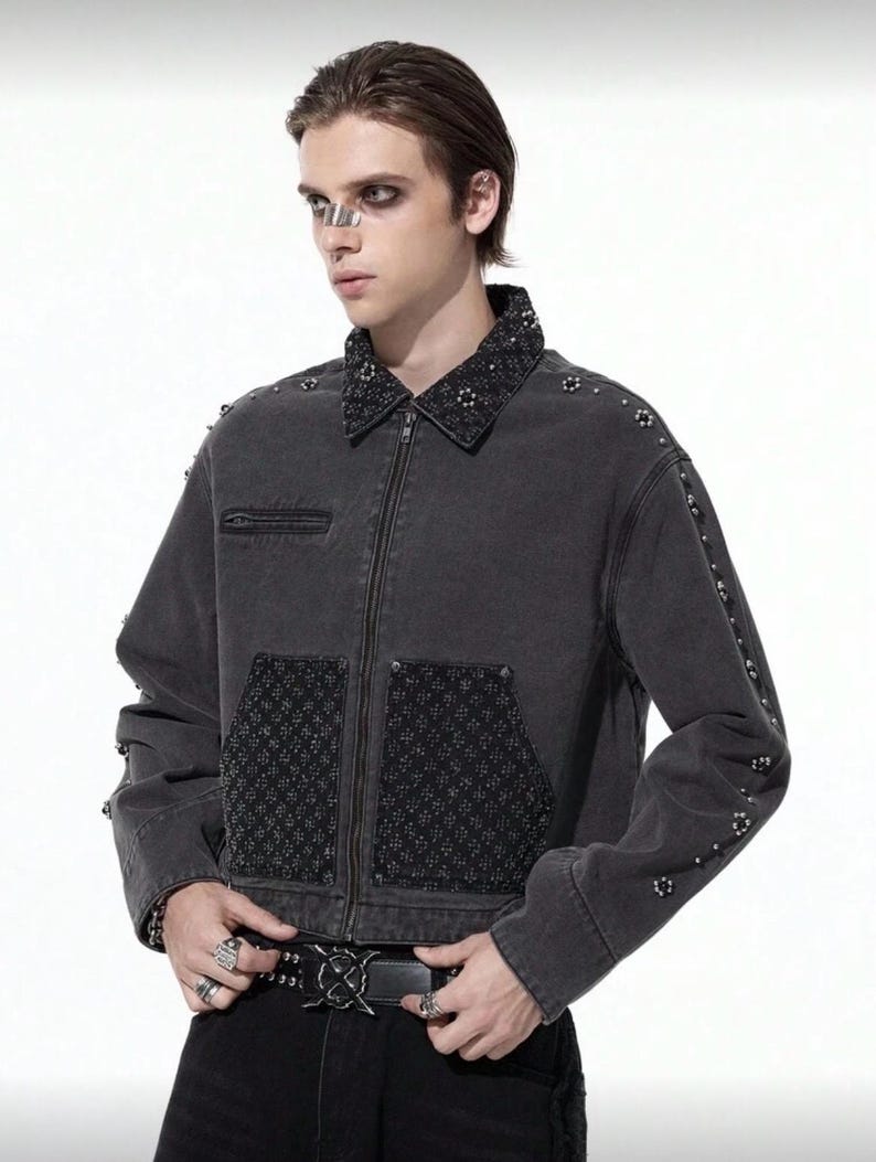 May include: A dark gray denim jacket with a zip-up front, embellished with studs on the collar, sleeves, and pockets. The jacket features a textured pattern on the pockets and collar. The model is wearing a black belt with a decorative buckle.