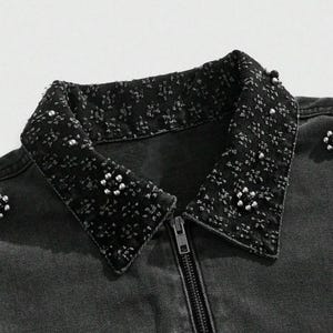 May include: Close-up of a black denim jacket with a decorative collar. The collar is embellished with small, silver-colored beads and a textured pattern. The jacket has a full-length zipper and a classic collar design.