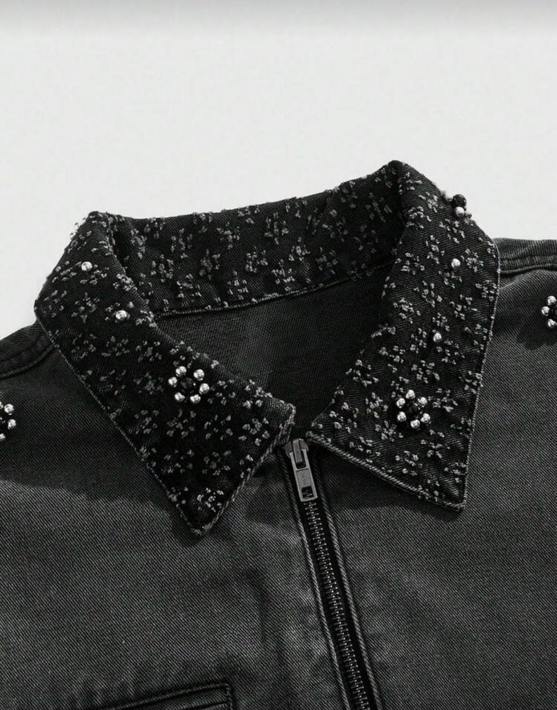 May include: Close-up of a black denim jacket with a decorative collar. The collar is embellished with small, silver-colored beads and a textured pattern. The jacket has a full-length zipper and a classic collar design.