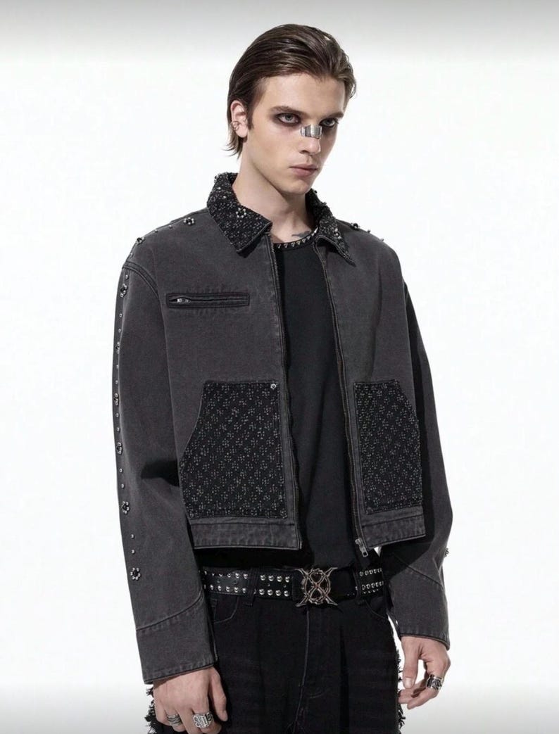 May include: A dark gray denim jacket with a studded collar, pocket accents, and sleeve details. The jacket features a zipper closure and is paired with a black belt with a decorative buckle. The jacket is part of a fashion ensemble.