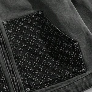May include: Close-up of a dark gray denim jacket with a black pocket featuring a textured floral pattern. The jacket has a silver zipper and silver rivets. The fabric appears to be pre-washed.