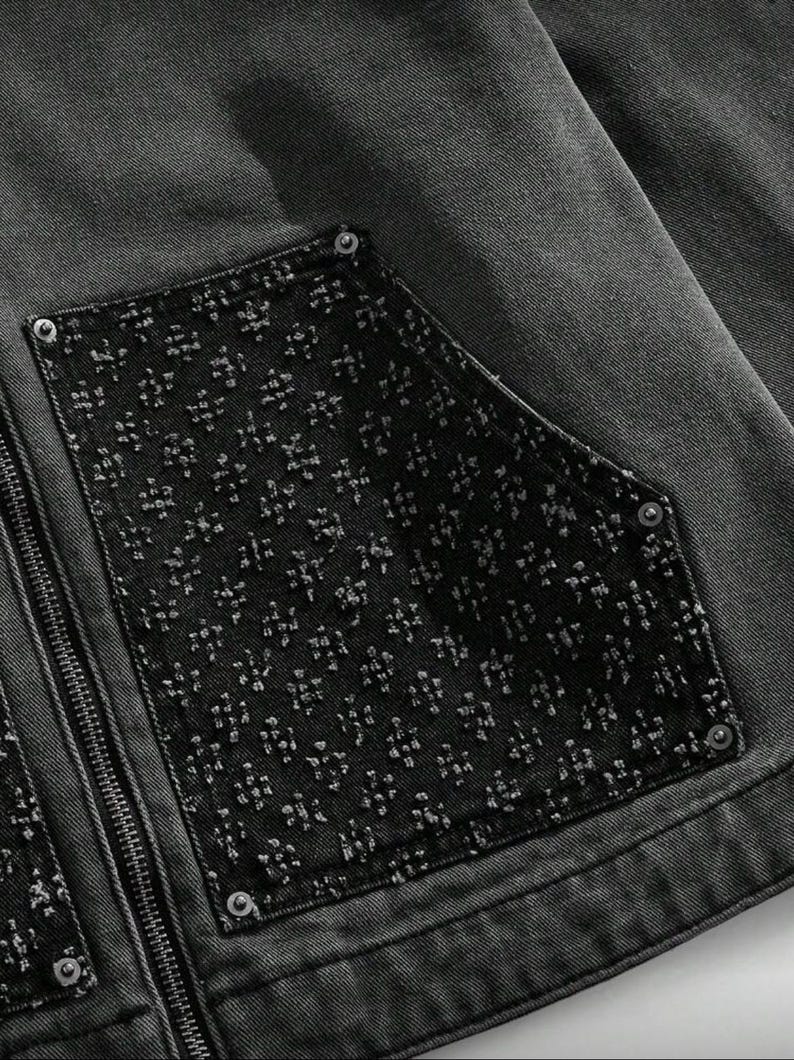 May include: Close-up of a dark gray denim jacket with a black pocket featuring a textured floral pattern. The jacket has a silver zipper and silver rivets. The fabric appears to be pre-washed.