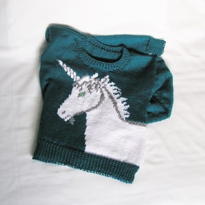 May include: Teal blue knitted sweater with a white unicorn design. The unicorn has a silver horn and green eyes.