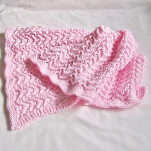 May include: A soft, pink knitted baby blanket with a textured, zig-zag pattern. The blanket is folded, showing the intricate stitch work and a ruffled edge. The blanket is made of a soft yarn, perfect for a baby.