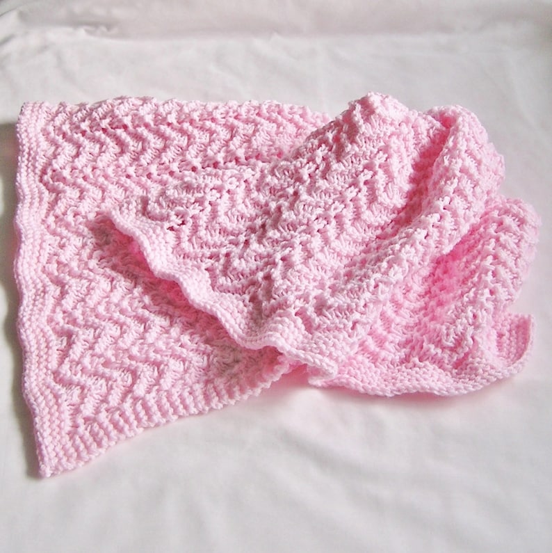 May include: A soft, pink knitted baby blanket with a textured, zig-zag pattern. The blanket is folded, showing the intricate stitch work and a ruffled edge. The blanket is made of a soft yarn, perfect for a baby.