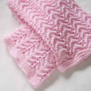 May include: A soft, light pink knitted blanket with a textured, open weave pattern. The blanket is folded, showcasing the intricate stitch work and the delicate color. The edges are finished with a neat, ribbed border, adding a touch of elegance.