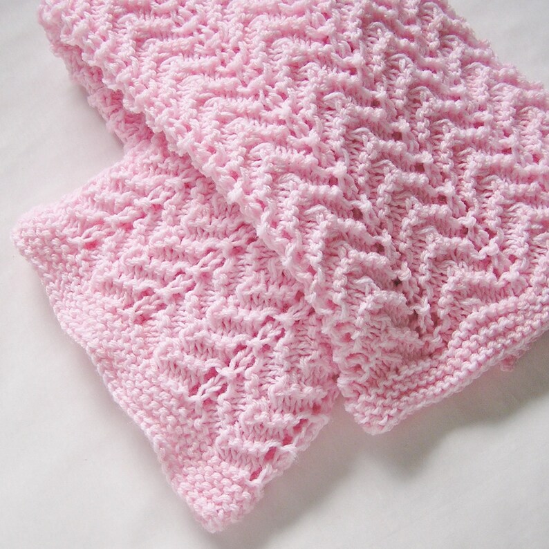 May include: A soft, light pink knitted blanket with a textured, open weave pattern. The blanket is folded, showcasing the intricate stitch work and the delicate color. The edges are finished with a neat, ribbed border, adding a touch of elegance.