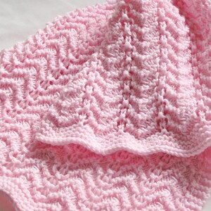 May include: A close-up of a soft, light pink crocheted blanket. The blanket features a textured, zig-zag pattern throughout. The edges are finished with a scalloped border. The blanket appears to be made of a soft yarn.