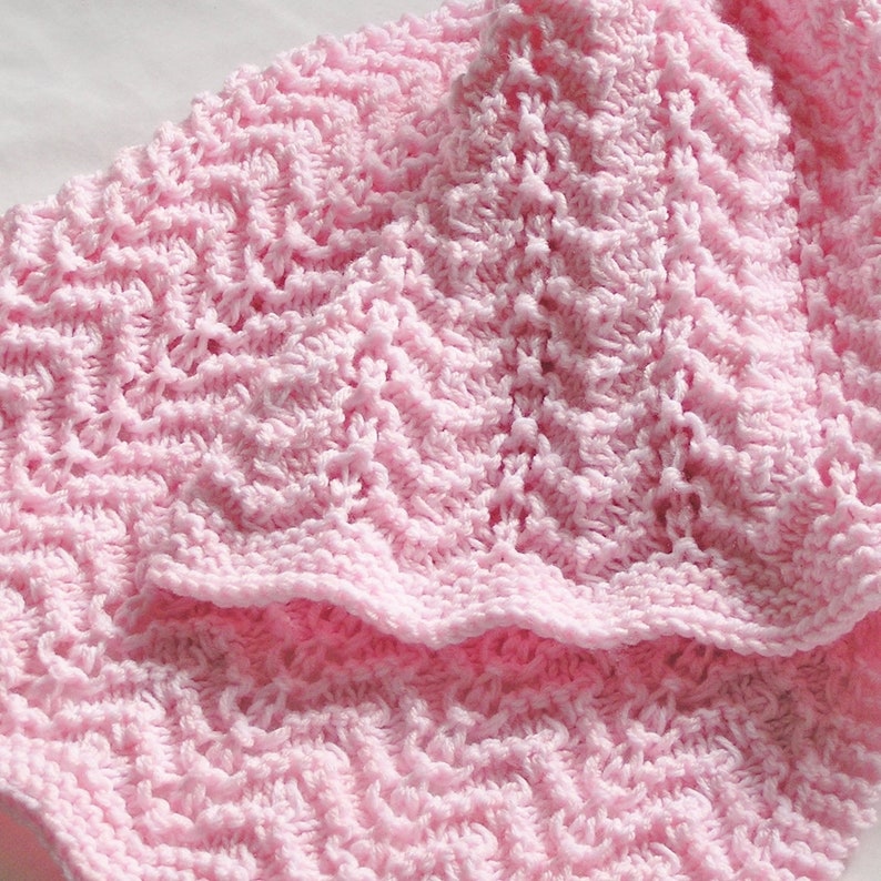 May include: A close-up of a soft, light pink crocheted blanket. The blanket features a textured, zig-zag pattern throughout. The edges are finished with a scalloped border. The blanket appears to be made of a soft yarn.