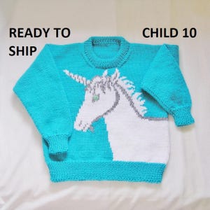 May include: A turquoise blue knitted sweater with a white unicorn design. The sweater has a crew neck and long sleeves. The text "CHILD 10" is visible in the top right corner of the image.