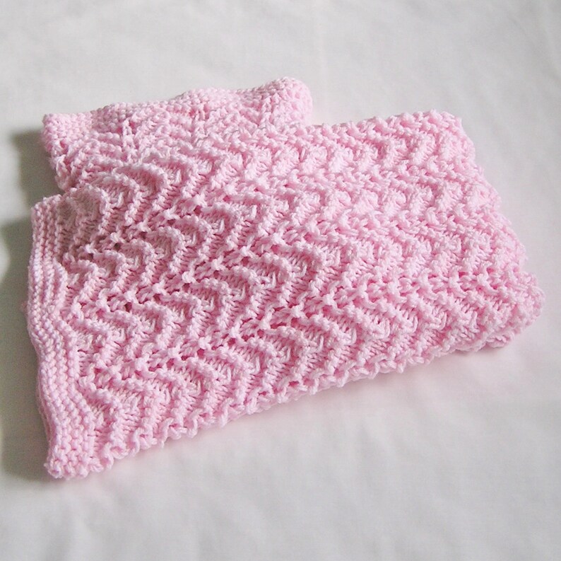 May include: A pair of light pink knitted baby blankets. The blankets feature a textured, zig-zag pattern. The soft, pastel color and intricate knit design suggest a handmade or artisanal quality, suitable for a nursery or as a baby gift.