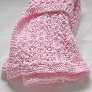 May include: A soft, light pink knitted baby blanket. The blanket features a textured pattern with a detailed stitch design. The edges are neatly finished, and the blanket is folded, showcasing its cozy and delicate appearance. The blanket is made of a soft yarn.