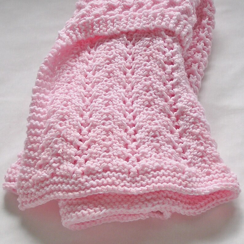May include: A soft, light pink knitted baby blanket. The blanket features a textured pattern with a detailed stitch design. The edges are neatly finished, and the blanket is folded, showcasing its cozy and delicate appearance. The blanket is made of a soft yarn.