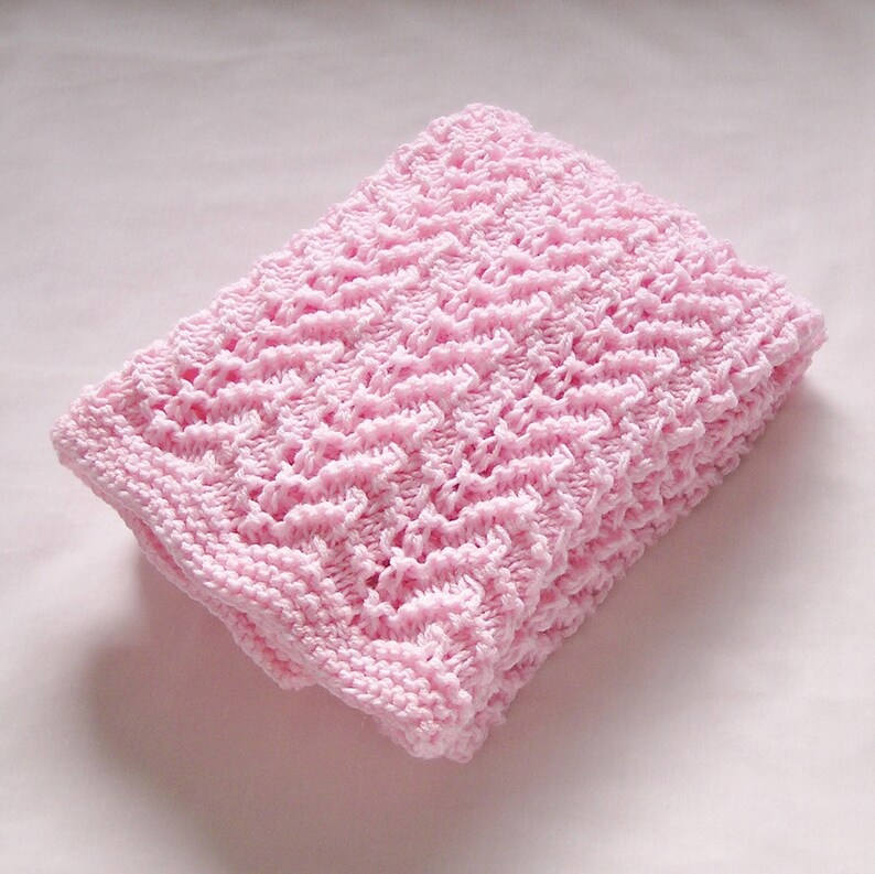 May include: A folded, light pink knitted blanket. The blanket features a textured, open weave pattern and a solid border. The soft pink color and delicate knit suggest a baby blanket or a decorative throw.