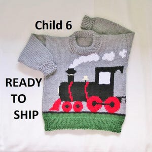 May include: A gray knitted sweater with a train design. The train is black with red wheels and a white smoke cloud. The sweater has a green bottom hem and the text "Child 6" and "READY TO SHIP".