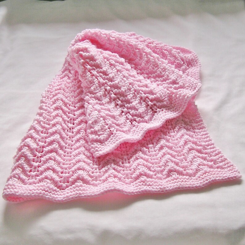May include: A soft, light pink knitted baby blanket with a textured chevron pattern. The blanket has a scalloped edge and is folded, showcasing the intricate stitch work. The blanket is made of a soft yarn, perfect for a baby.