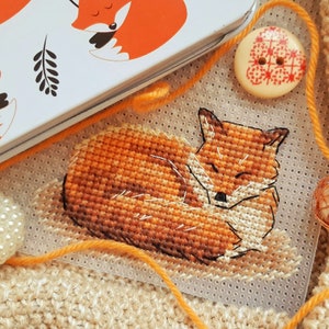 May include: A close-up of a cross-stitch embroidery of a sleeping orange fox. The fox is curled up on a white background with a grid pattern.