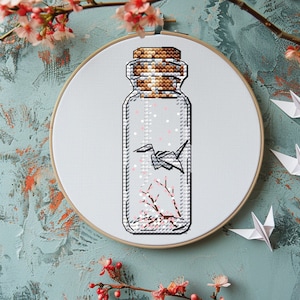 Op de afbeelding: Een witte borduurring met een glazen fles erin. De fles bevat een kleine origami-kraanvogel en roze kersenbloesems. De fles heeft een kurken stop.