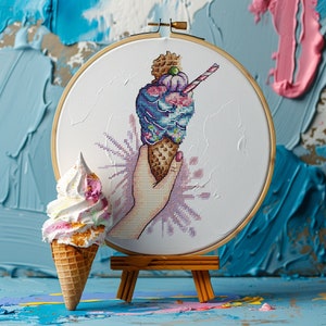 Peut inclure: Une main tenant un cornet de glace coloré avec un tourbillon rose, bleu et blanc. La glace est dans un cerceau de broderie en bois.