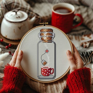 Può includere: Un primo piano di un telaio da ricamo a punto croce con un disegno di un barattolo di vetro con una bustina di tè all'interno e una tazza rossa e bianca a pois sul fondo. Il barattolo ha un tappo di sughero e la bustina di tè è appesa alla parte superiore del barattolo.