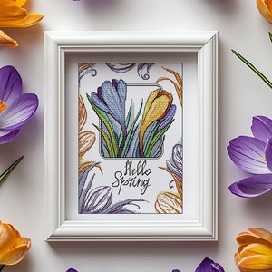 May include: A white frame containing a cross-stitch design of purple and yellow crocuses with green leaves. The text "Hello Spring" is written in black cursive lettering.