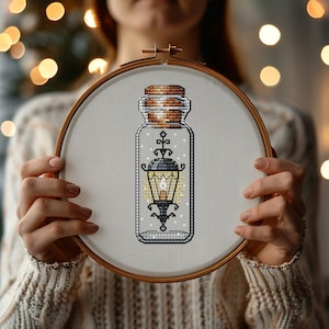 May include: A cross-stitch embroidery hoop with a white background and a clear glass jar with a cork lid. Inside the jar is a black and white cross-stitch design of a street lamp with a lit candle.