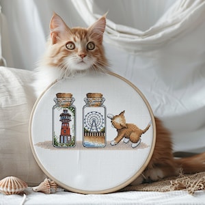 Puede incluir: Un aro de bordado blanco con un diseño de punto de cruz de dos frascos de vidrio que contienen un faro y una noria. Un gato jengibre está sentado junto al aro, mirando los frascos.