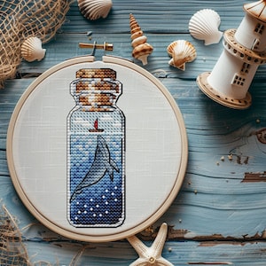 以下が含まれることがあります： 青いガラス瓶の中にクジラと小さな帆船が入ったクロスステッチ刺繍のフープ。瓶にはコルク栓が付いており、白い布で囲まれています。