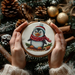 Puede incluir: Un primer plano de un aro de bordado hecho a mano con un fondo blanco y un lindo pingüino de dibujos animados que lleva una bufanda roja y blanca. El pingüino está sentado en un trineo con acebo verde y bayas rojas.