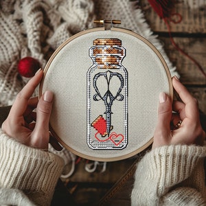 Peut inclure: Un cerceau de broderie blanc avec un motif de point de croix représentant un bocal en verre contenant une paire de ciseaux, une bobine de fil et un cœur. Le bocal a un couvercle en liège.