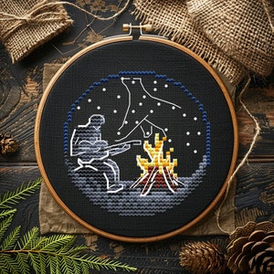 May include: Cross-stitch embroidery in a wooden hoop. The design features a person playing a guitar by a campfire under a starry sky. The color palette includes black, white, gray, yellow, and brown.