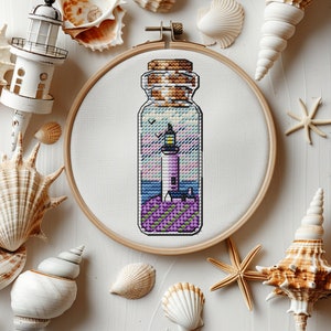 Puede incluir: Un aro de bordado de punto de cruz con fondo blanco y una botella de vidrio con tapón de corcho. La botella contiene una imagen de punto de cruz de un faro en una isla morada con cielo azul y mar.