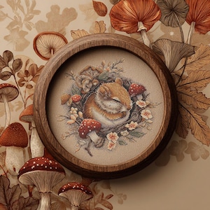 May include: A framed cross-stitch featuring a sleeping squirrel nestled among mushrooms and flowers. The artwork is set in a round, wooden frame. The color palette includes shades of brown, orange, and red, creating a cozy, autumnal theme.