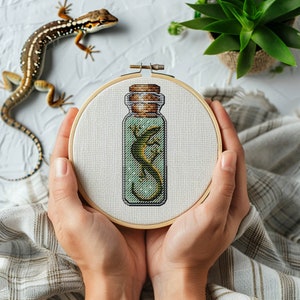 Könnte beinhalten: Nahaufnahme einer Hand, die einen kleinen, runden Stickrahmen mit einem Kreuzstich-Motiv eines grünen Leguan in einer Glasflasche hält. Die Flasche hat einen Korkverschluss und der Leguan ist darin eingerollt.