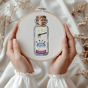 May include: A glass jar with a cork lid containing a colorful pincushion and a dragonfly. The jar is on a white fabric background.
