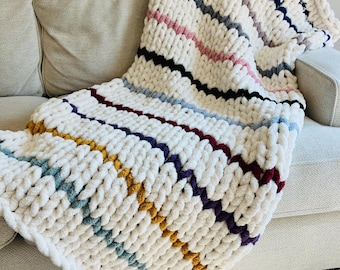 May include: A chunky knit throw blanket with a white base and colorful stripes in shades of blue, purple, pink, yellow, and gray. The blanket is draped over a beige couch.