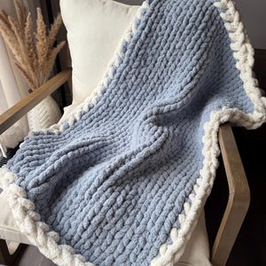 Può includere: Una morbida coperta di maglia blu con un bordo intrecciato bianco. La coperta è drappeggiata su una sedia.