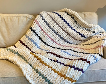 May include: A chunky knit throw blanket with a white base and colourful stripes in shades of blue, grey, pink, red, purple, and yellow. The blanket is draped over a light beige sofa.