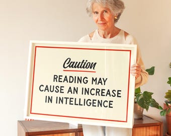 May include: A framed sign with the word "Caution" in cursive above the text "Reading may cause an increase in intelligence" in black. The sign has a cream background with a red border. The sign is held by a person.