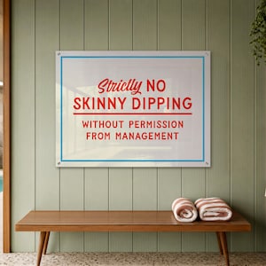 Op de afbeelding: Een rechthoekig wandbord met een witte achtergrond, een dunne blauwe rand en rode tekst die luidt: Strictly NO SKINNY DIPPING WITHOUT PERMISSION FROM MANAGEMENT. Het bord is bevestigd met vier zilverkleurige bevestigingsmiddelen op de hoeken.