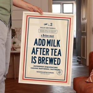 May include: A framed poster with a cream background, red and blue borders, and dark blue text that reads: No. 2 RULES BRITANNIA OFFICIAL DIRECTIVES FROM THE MINISTRY OF BRITISH IDENTITY a Briton must ADD MILK AFTER TEA IS BREWED.