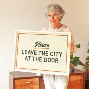 May include: A framed sign with the text "Please Leave the City at the Door" in a black, stylized font. The sign has a cream-colored background with a brown border and is held by a person.