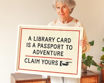 May include: A framed sign with the text "A LIBRARY CARD IS A PASSPORT TO ADVENTURE CLAIM YOURS" in black font on a cream background, with red border. A hand points to the text. The sign is held by a person.