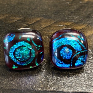 May include: A pair of square-shaped glass earrings with a dark red base and a swirling blue and green design.